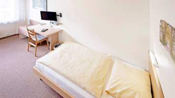 Una habitación pequeña y ordenada con una cama individual, un escritorio con computadora y silla, y un ventanal con cortinas.