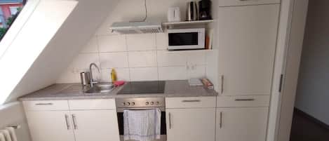 A compact kitchen with a gas stove, microwave, and a small oven. There is a window providing natural light, and the kitchen has white cabinets and a tiled backsplash.