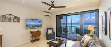 A living room with a sofa, coffee table, and a chair. There is a TV on the wall and a ceiling fan.