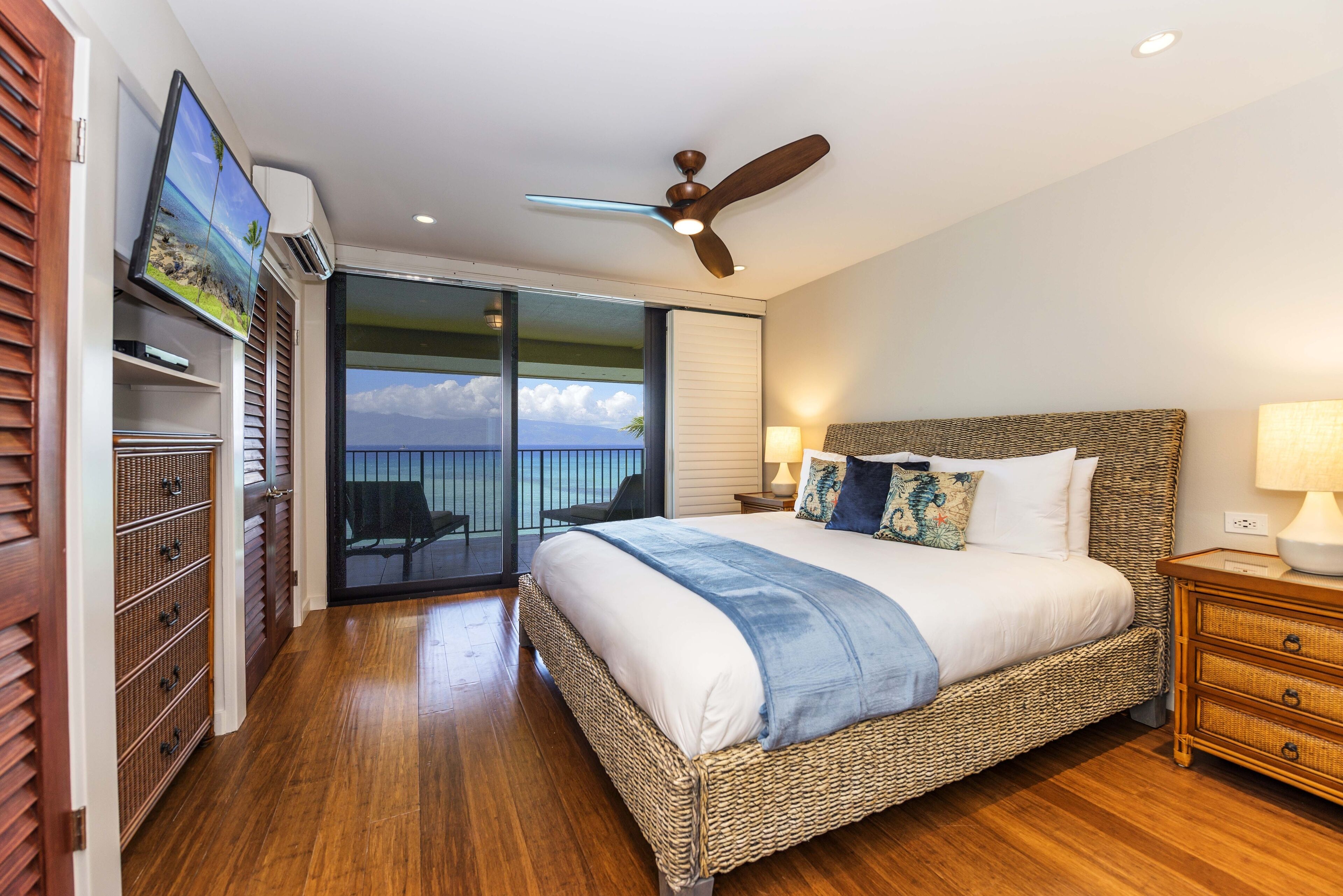 A bedroom with a large bed, a ceiling fan, a TV mounted on the wall, and a balcony with a view of the sky.