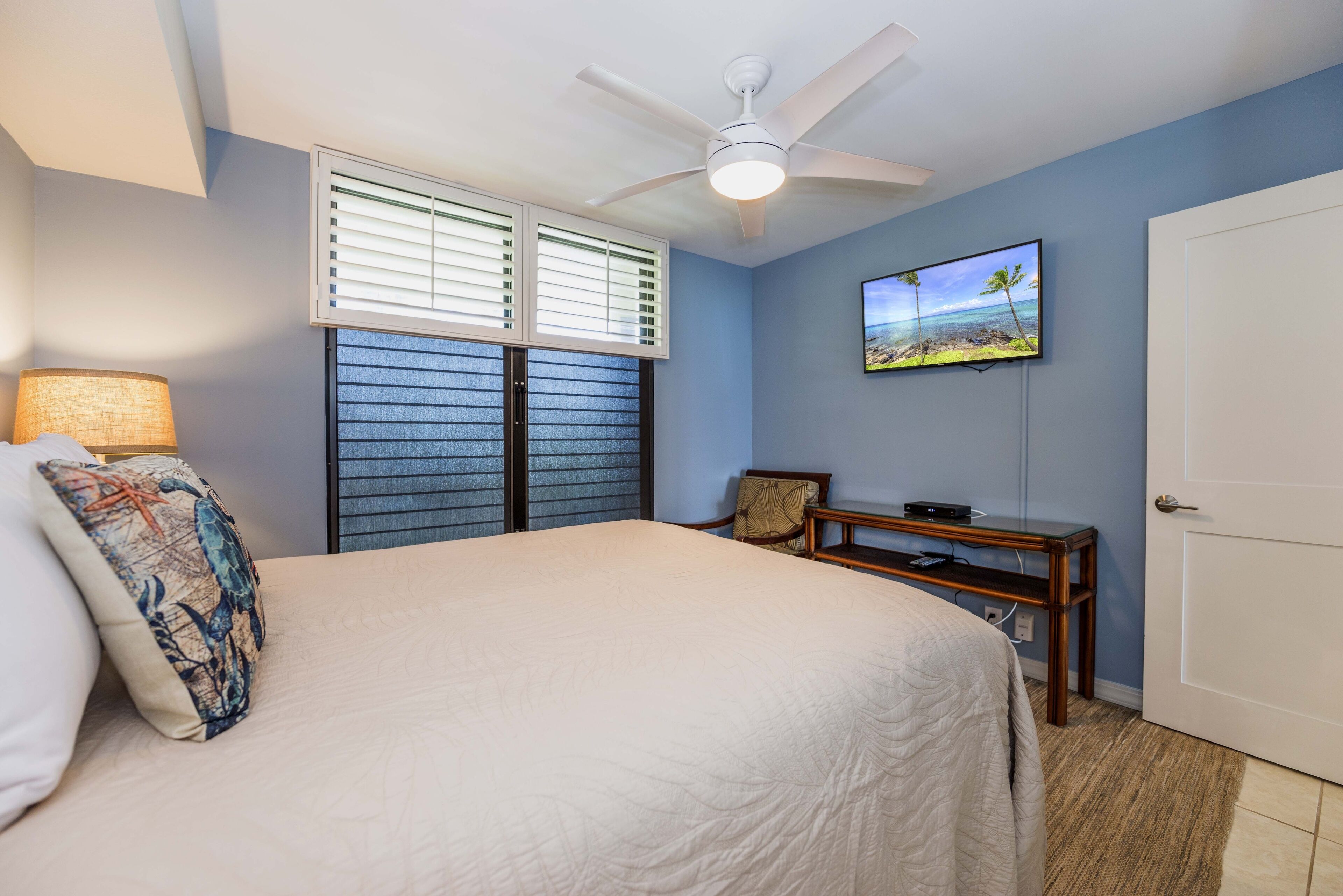 A bedroom with a bed, a ceiling fan, a TV on the wall, and a door.
