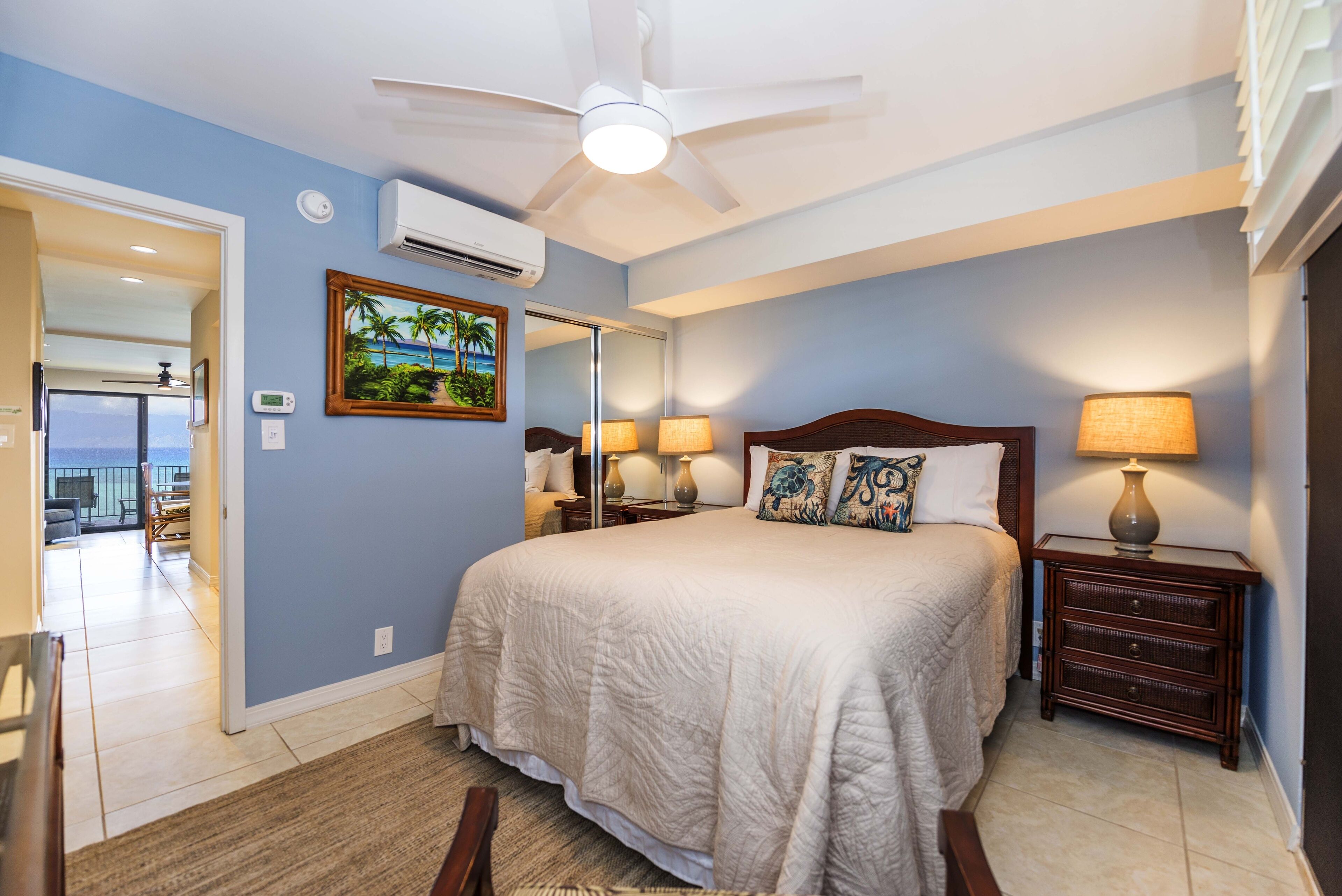 Une chambre à coucher comprenant un lit, des tables de chevet, un ventilateur de plafond, un appareil de climatisation et un tableau représentant un paysage tropical.