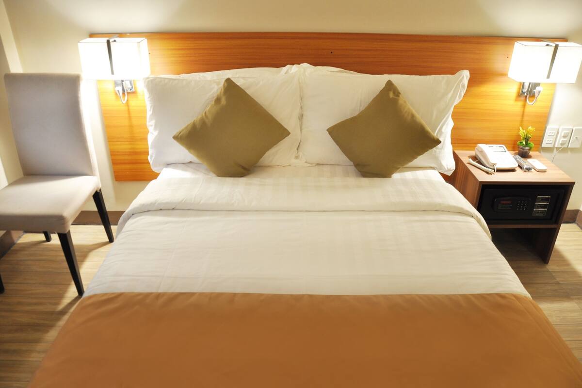 A hotel room with a bed, two bedside lamps, a nightstand with a phone and remote, and a chair.