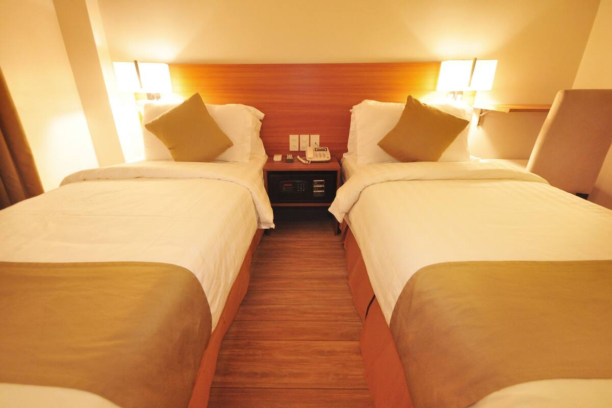 A hotel room with two single beds, a wooden headboard, bedside lamps, a nightstand with a phone and a lamp, and a wooden floor.