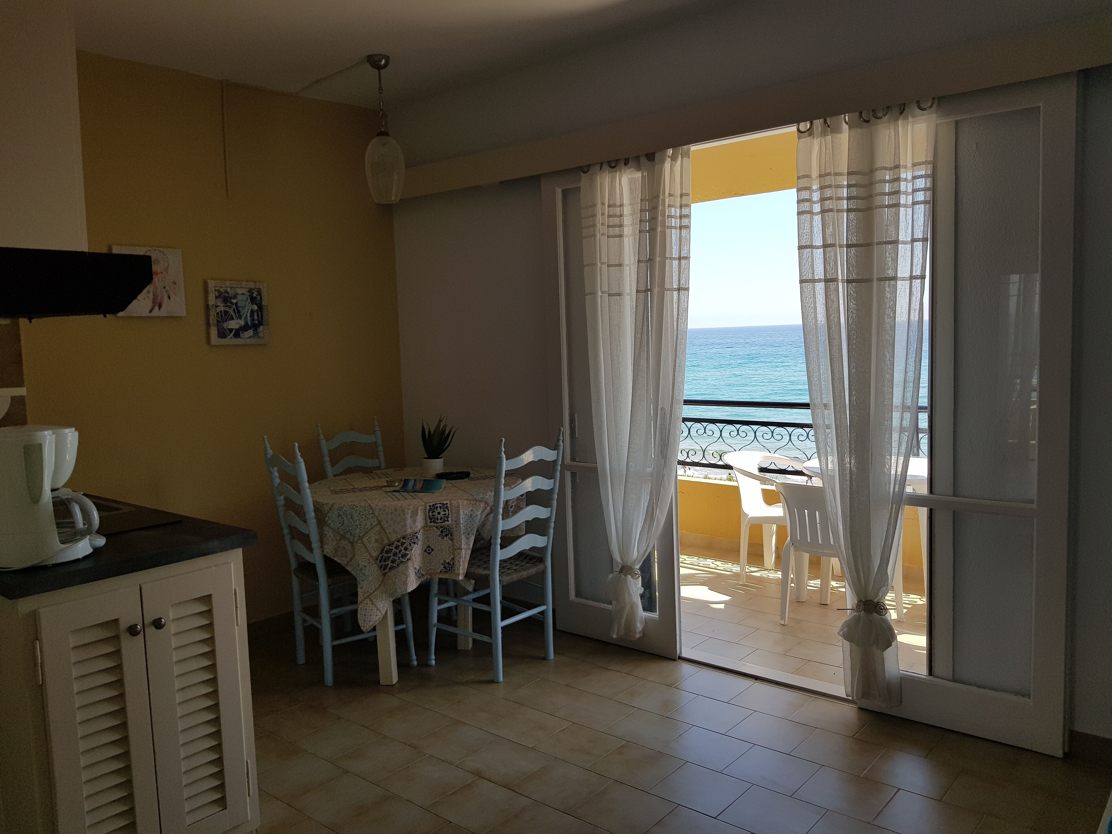 A room with a dining table, chairs, a coffee maker, and a view of the sea through large windows.