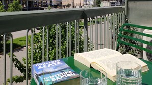 Un balcón con un libro y gafas sobre una mesa verde, con vista a un paisaje urbano que incluye edificios y una colina.