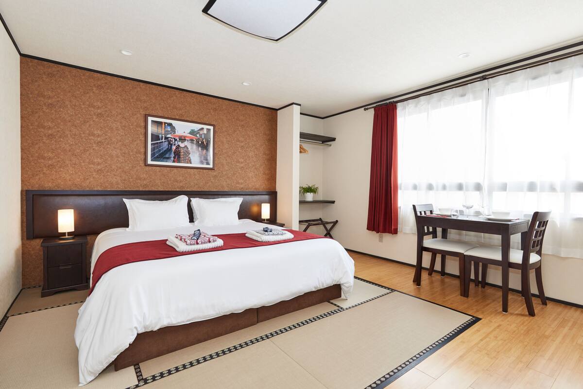 A hotel room with a large bed, a desk with chairs, a window with curtains, and a picture on the wall.