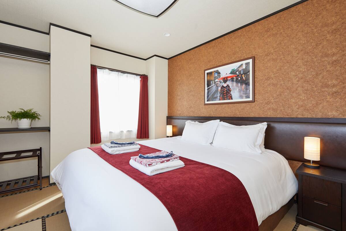 A hotel room with a large bed, a nightstand with a lamp, a window with red curtains, and a framed picture on the wall.