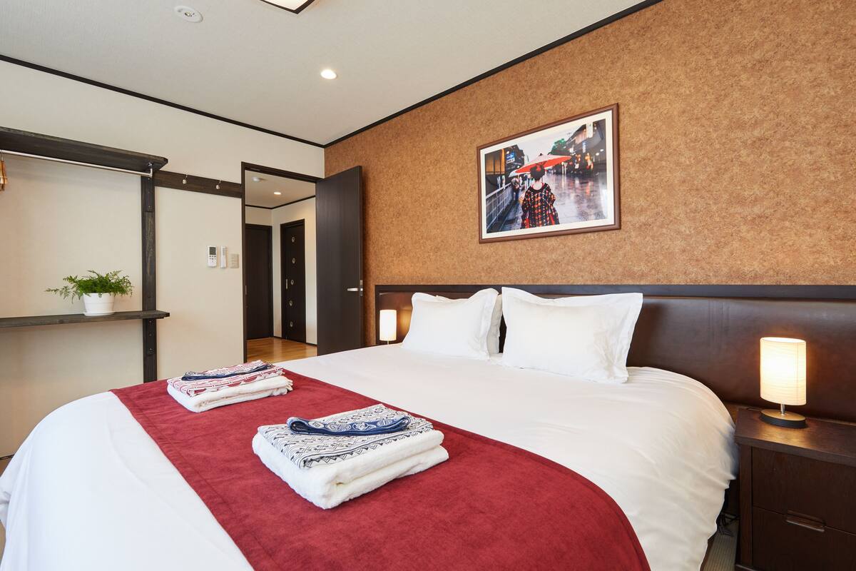 A hotel room with a large bed, a red throw blanket, neatly folded towels, a nightstand with a lamp, and a framed picture on the wall.