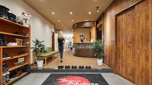 Un hall d’hôtel comprenant un comptoir d’accueil, des étagères à chaussures, un banc vert et un logo noir et rouge sur le sol.