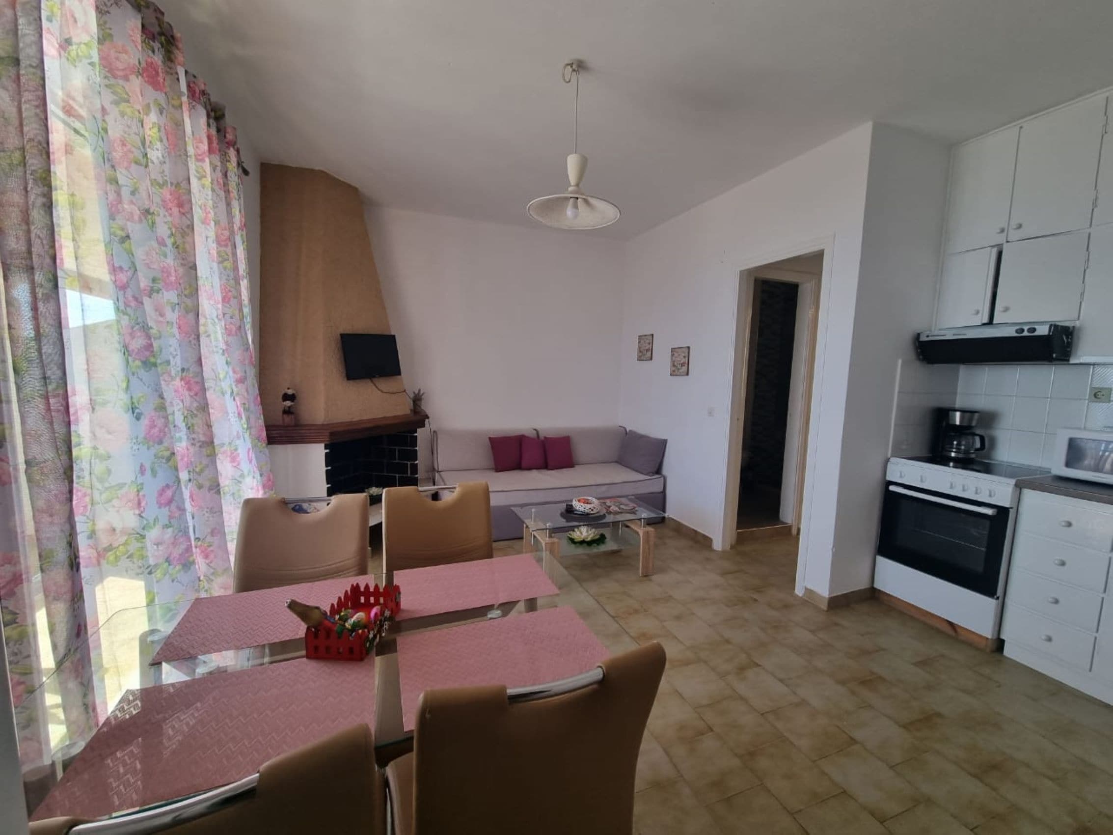 A living room with a sofa, a coffee table, and a TV. There is a dining area with a table and chairs. The kitchen has a stove and cabinets.