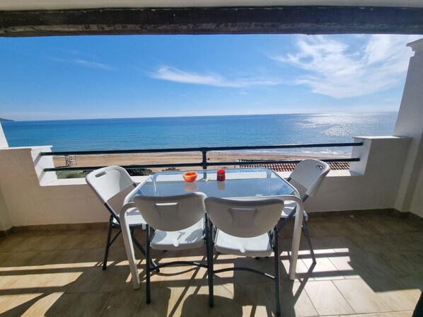 Un balcon doté d’une table en verre et de quatre chaises, offrant une vue sur une plage et la mer.