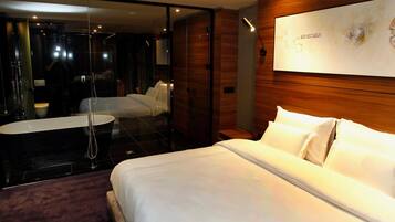 A modern hotel room with a large bed, a separate seating area, a glass-enclosed shower, and a wooden accent wall.