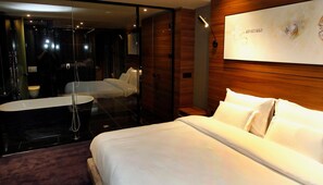 A modern hotel room with a large bed, a separate seating area, a glass-enclosed shower, and a wooden accent wall.