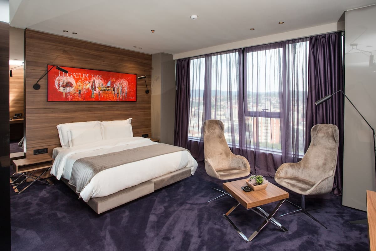 A modern hotel room with a large bed, two armchairs, a small table, a painting on the wall, and a view of the city through large windows.