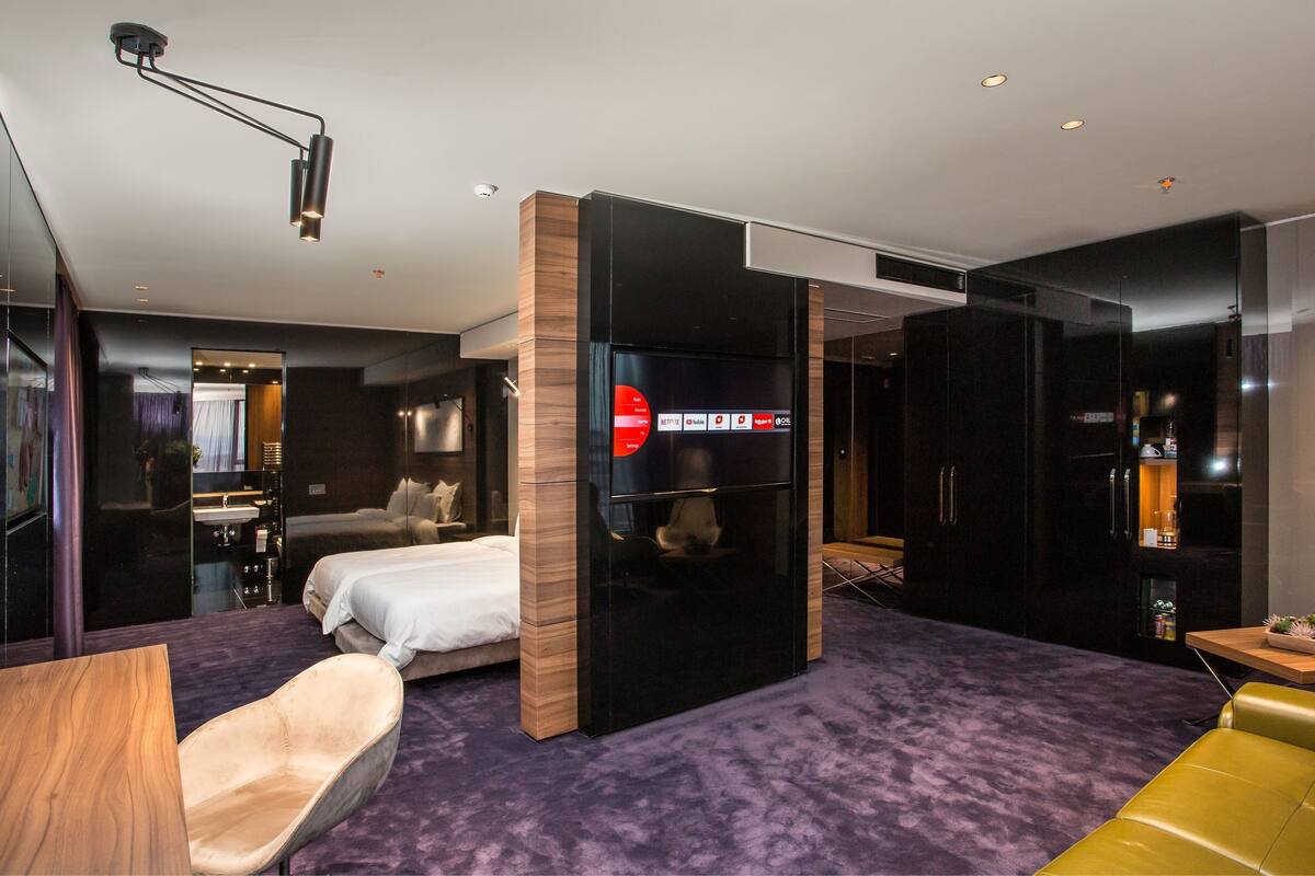 A modern hotel room with a bed, a seating area with a yellow sofa, a small table, and a TV mounted on a wooden partition.
