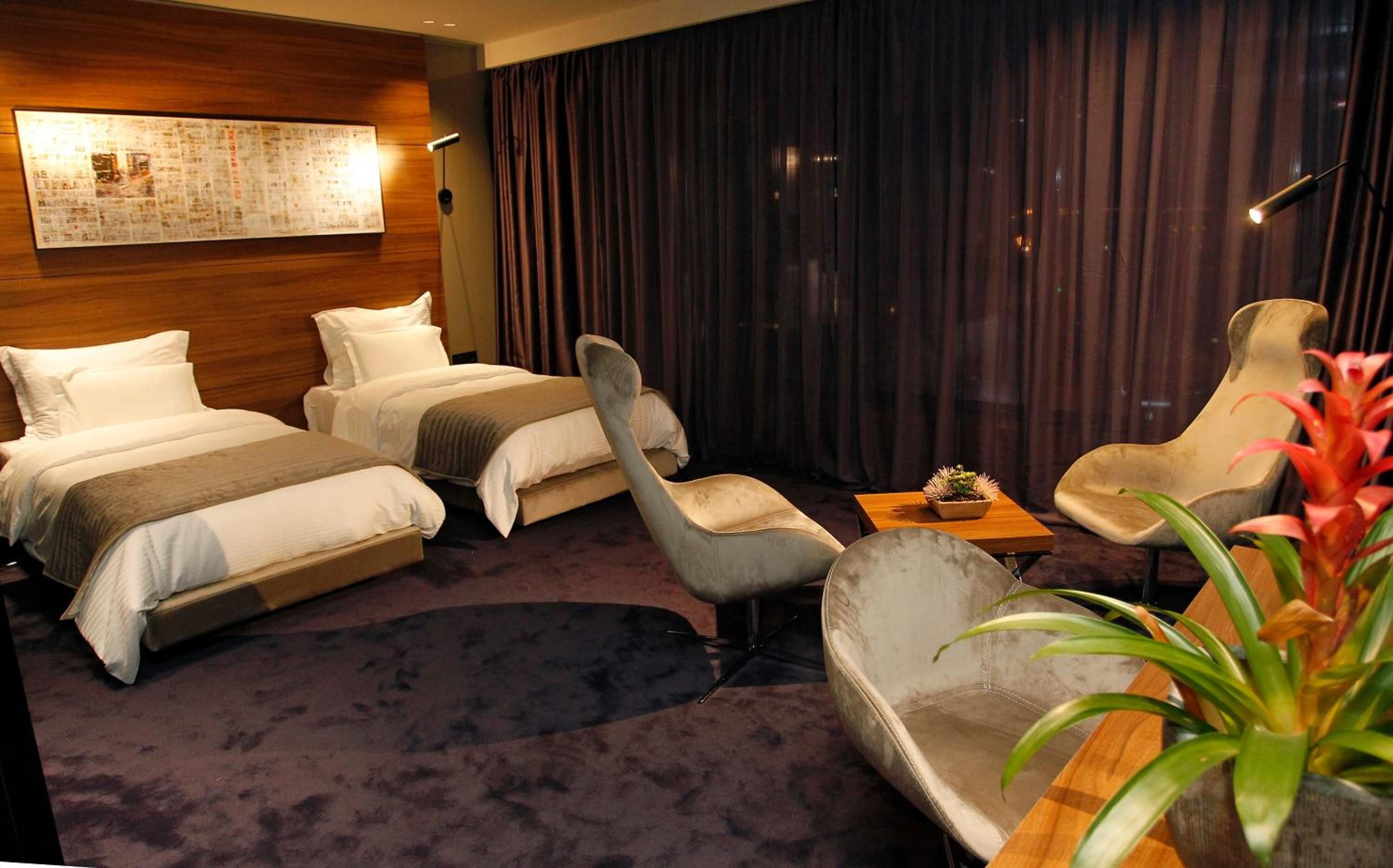 A hotel room with two beds, a grey armchair, a wooden side table, and a potted plant.