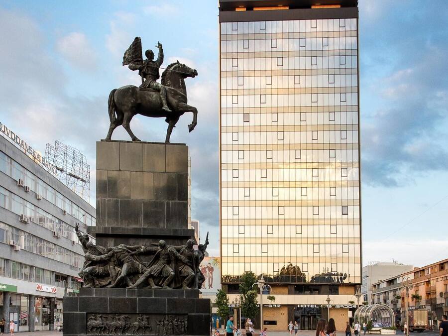 Een standbeeld van een paard en ruiter in een openbaar plein, met een modern gebouw op de achtergrond.