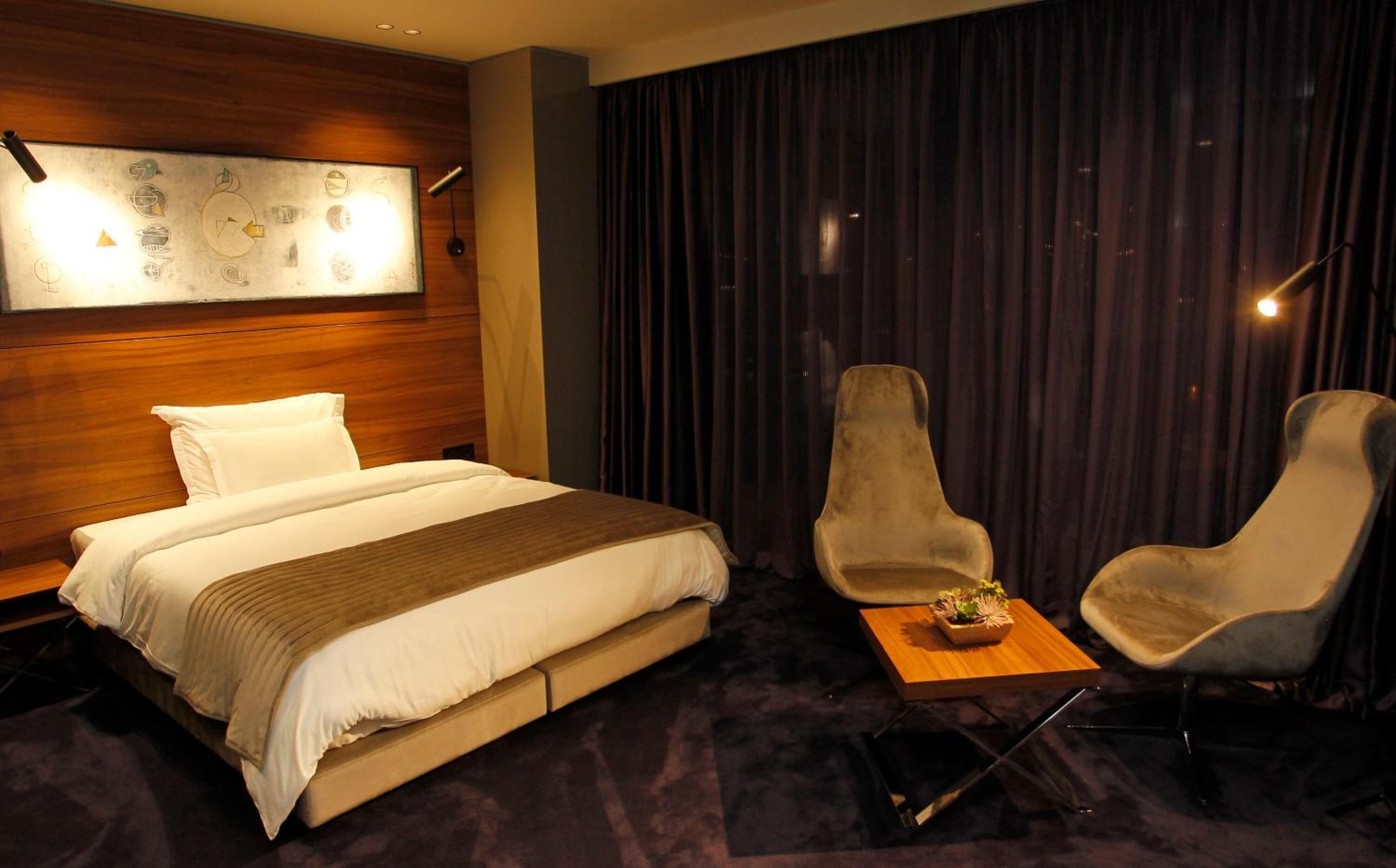 A hotel room with a bed, two armchairs, a small table with a plant, and a wall with abstract art.