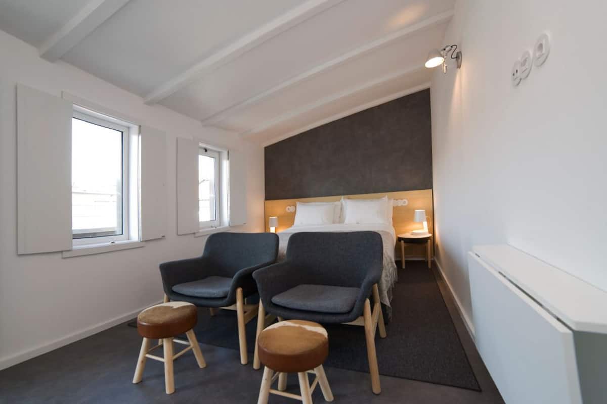A modern hotel room with a large bed, two armchairs, a small stool, and a nightstand.