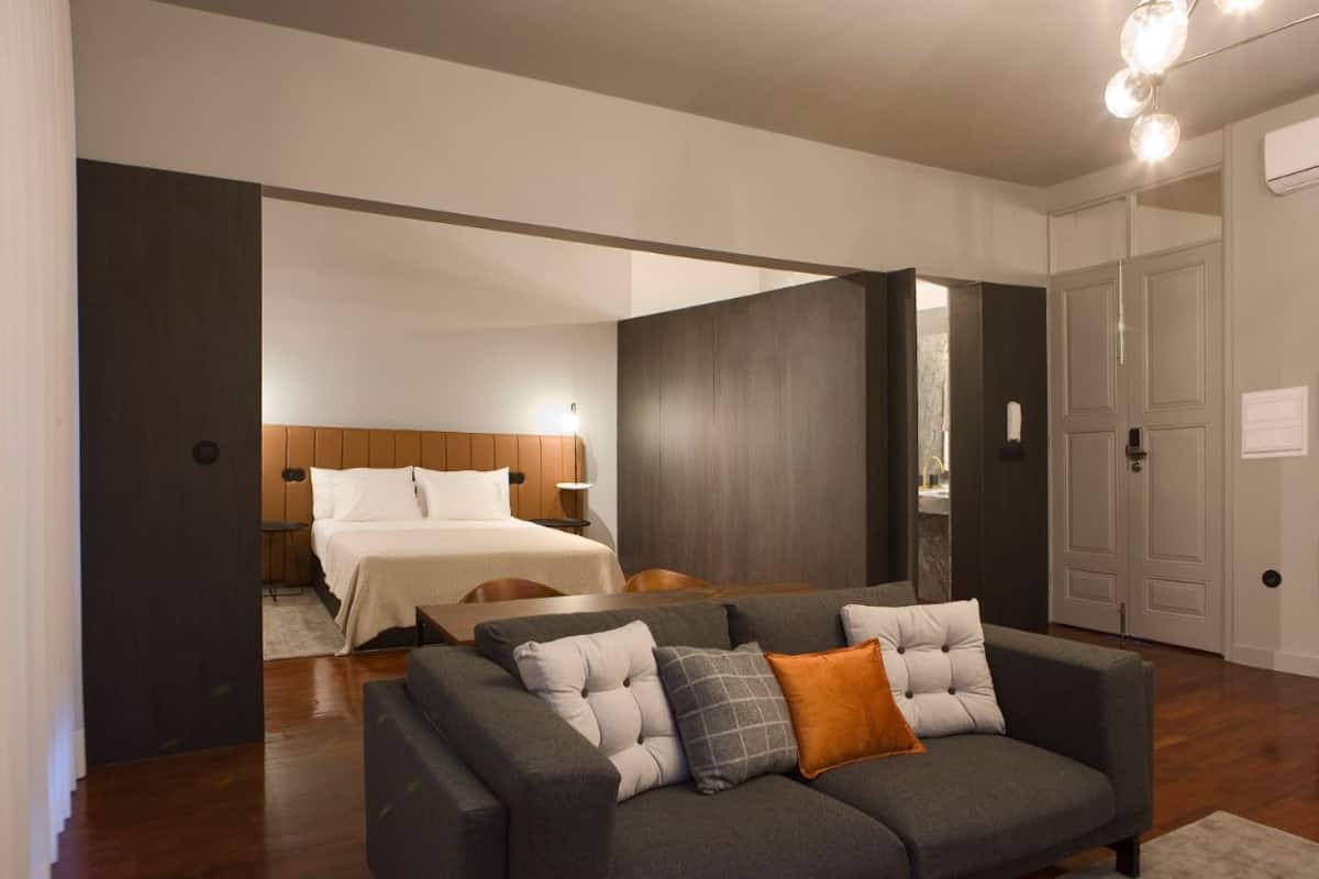 A modern hotel room with a large bed, a grey sofa, and a wooden floor.