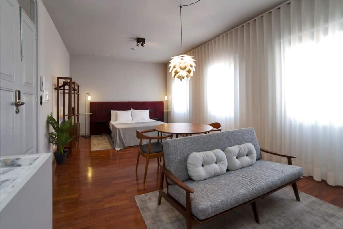 A modern hotel room with a wooden floor, a grey sofa, a round dining table, a bed with a red headboard, and a large window with sheer curtains.