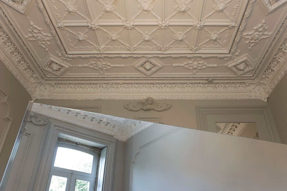 A room with a high, ornate ceiling featuring intricate plasterwork and a large window allowing natural light.