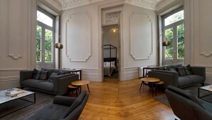 Une pièce spacieuse avec un plafond haut, des moulures décoratives et un lustre. On y trouve deux canapés et une table basse, et de grandes fenêtres offrent une vue sur un jardin.