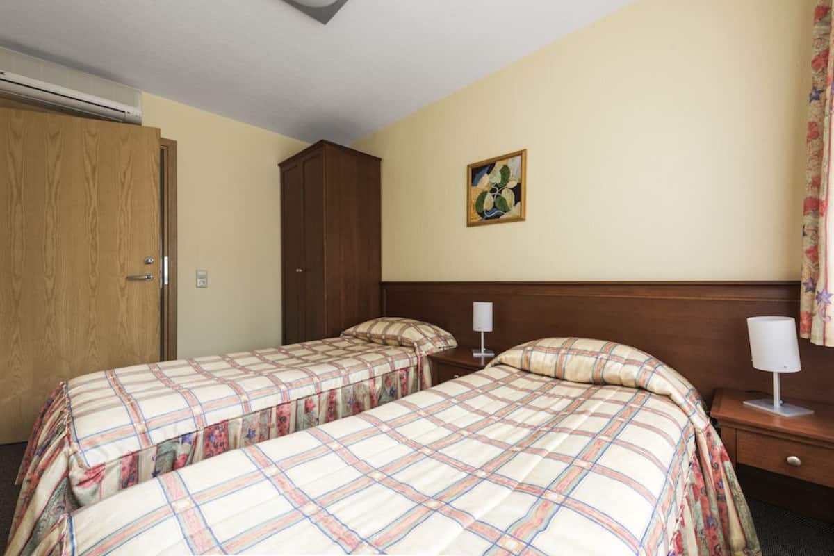 A hotel room with two beds, a wooden wardrobe, a painting on the wall, and a nightstand with a lamp.
