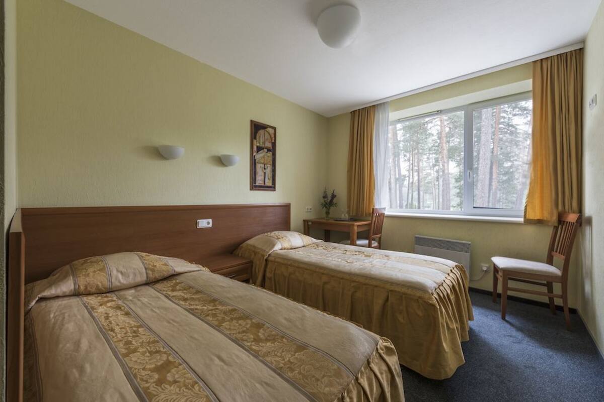A hotel room with two single beds, a desk with a chair, a window with curtains, and a painting on the wall.