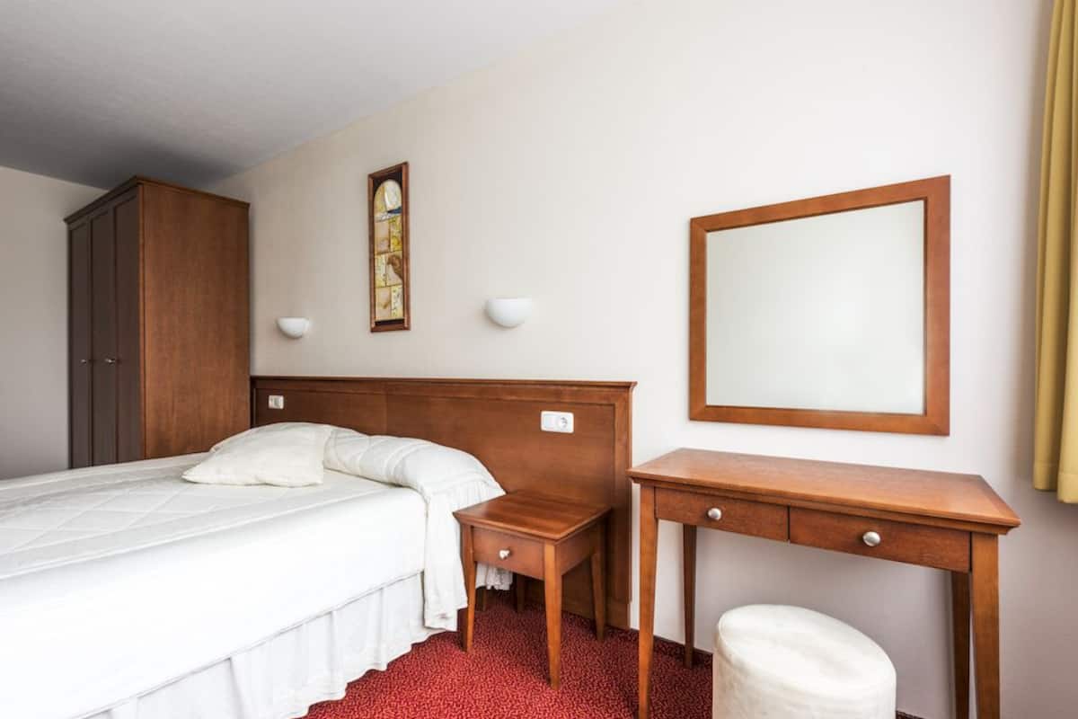 A hotel room with a bed, a wooden nightstand, a desk, a mirror, and a wardrobe.