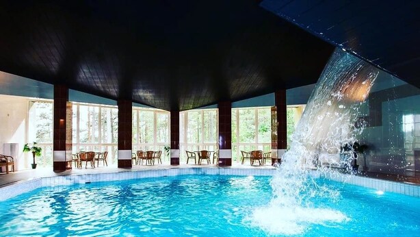 An indoor swimming pool with clear water, surrounded by wooden chairs and tables, and large windows allowing natural light. - ORO Dubingiai (Giraiciai)