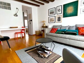 A living room with a grey sofa, red and green cushions, a coffee table, and a bookshelf. There are framed pictures on the wall and a small table with a lamp.