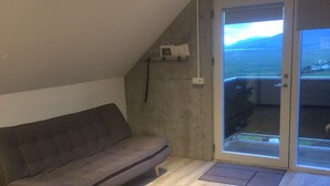 A room with a grey sofa, wooden flooring, a ceiling light, a sliding glass door with a view of the outdoors, and a small shelf with items.