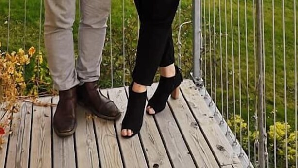 Two people standing on a wooden deck with a metal railing, overlooking a mountainous landscape.