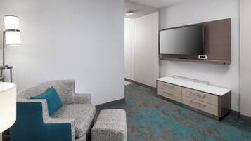 Una habitación de hotel moderna con sofá gris, cojines color esmeralda, un televisor de pantalla plana, una mesita de noche y una alfombra estampada.