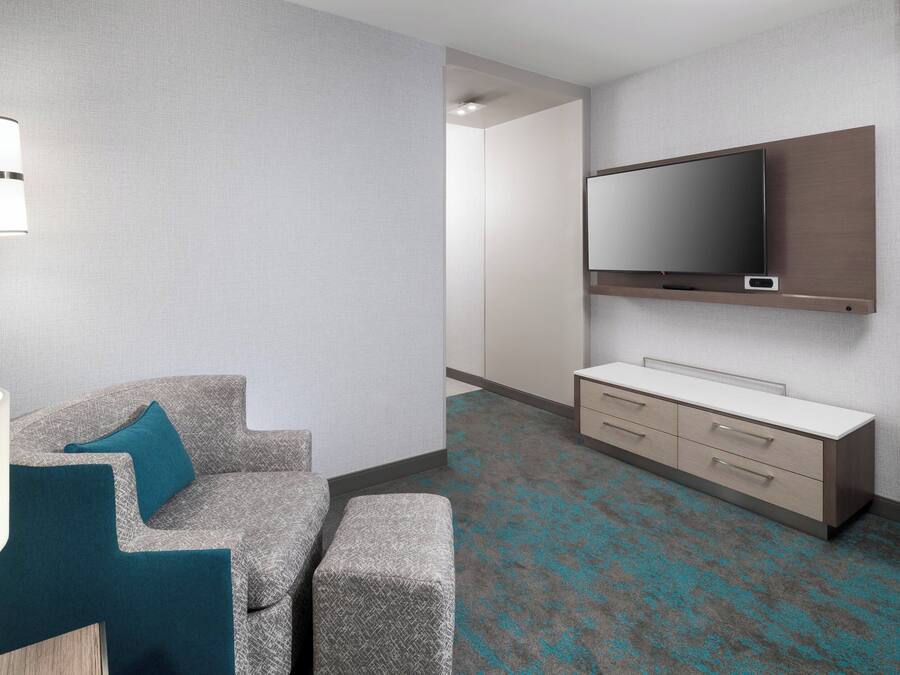 Una habitación de hotel moderna con sofá gris, cojines color esmeralda, un televisor de pantalla plana, una mesita de noche y una alfombra estampada.