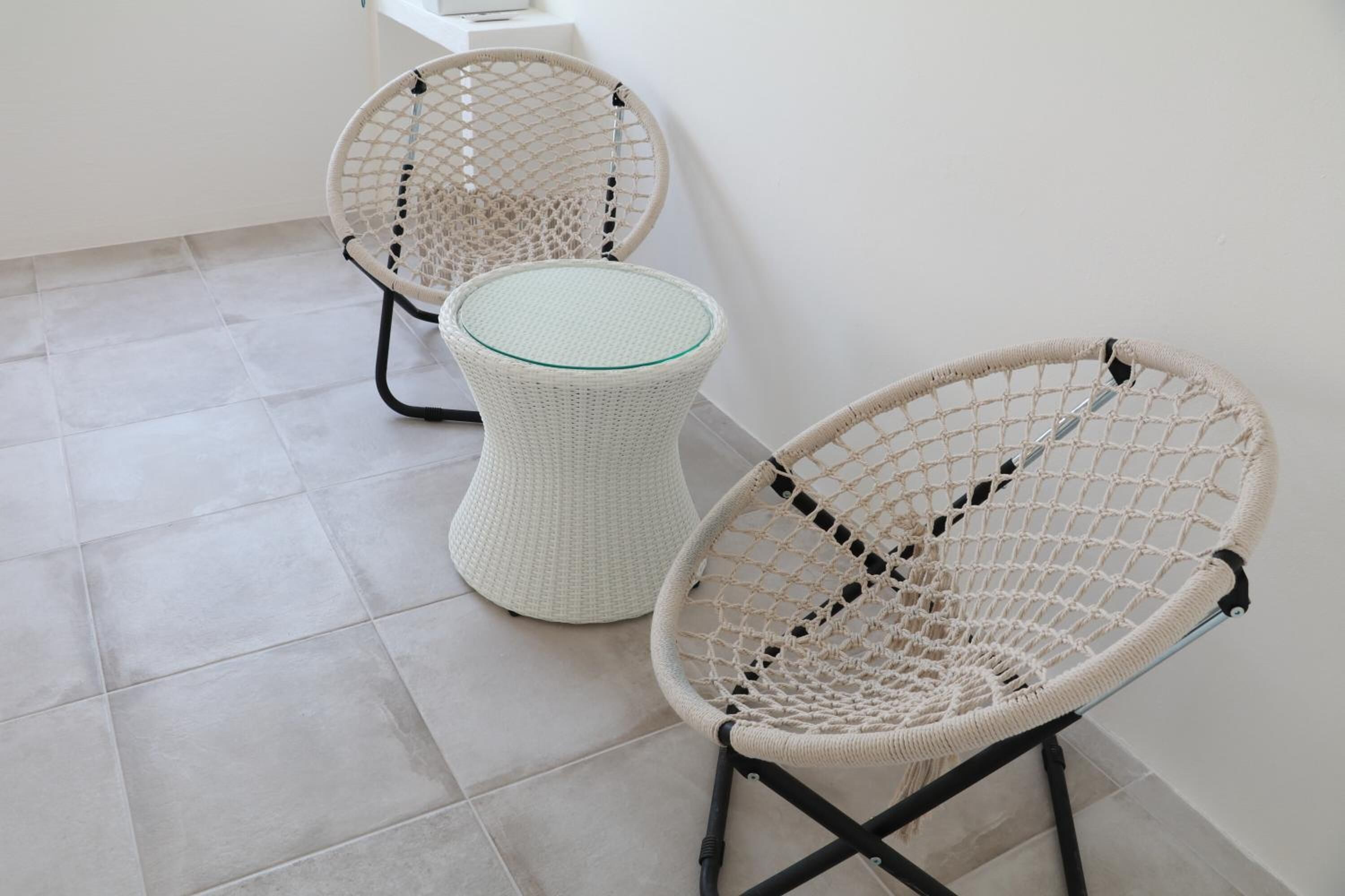 Deux fauteuils en osier avec des plateaux en verre, placés sur un sol carrelé, adossés à un mur uni.