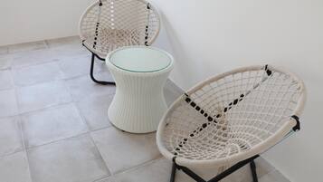 Deux fauteuils en osier avec des plateaux en verre, placés sur un sol carrelé, adossés à un mur uni.