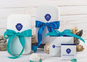 Drie cadeau dozen met Miura branding van Mykonos Resort, vastgebonden met turquoise linten, geplaatst op een houten oppervlak met schelpen.