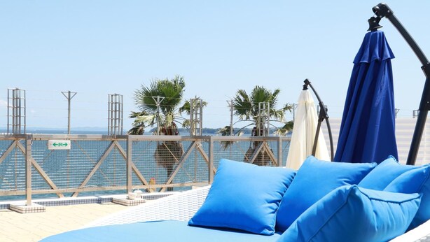 Un espace extérieur lumineux, aménagé pour se détendre, avec vue sur la mer, comprenant un fauteuil de jardin en osier blanc agrémenté de coussins bleus et d’un parasol bleu.