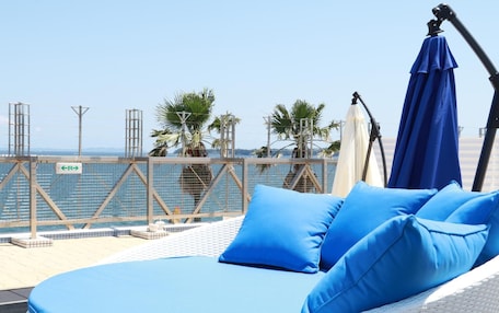 A sunlit outdoor seating area with a view of the sea, featuring a white wicker lounge chair with blue cushions and a blue umbrella.