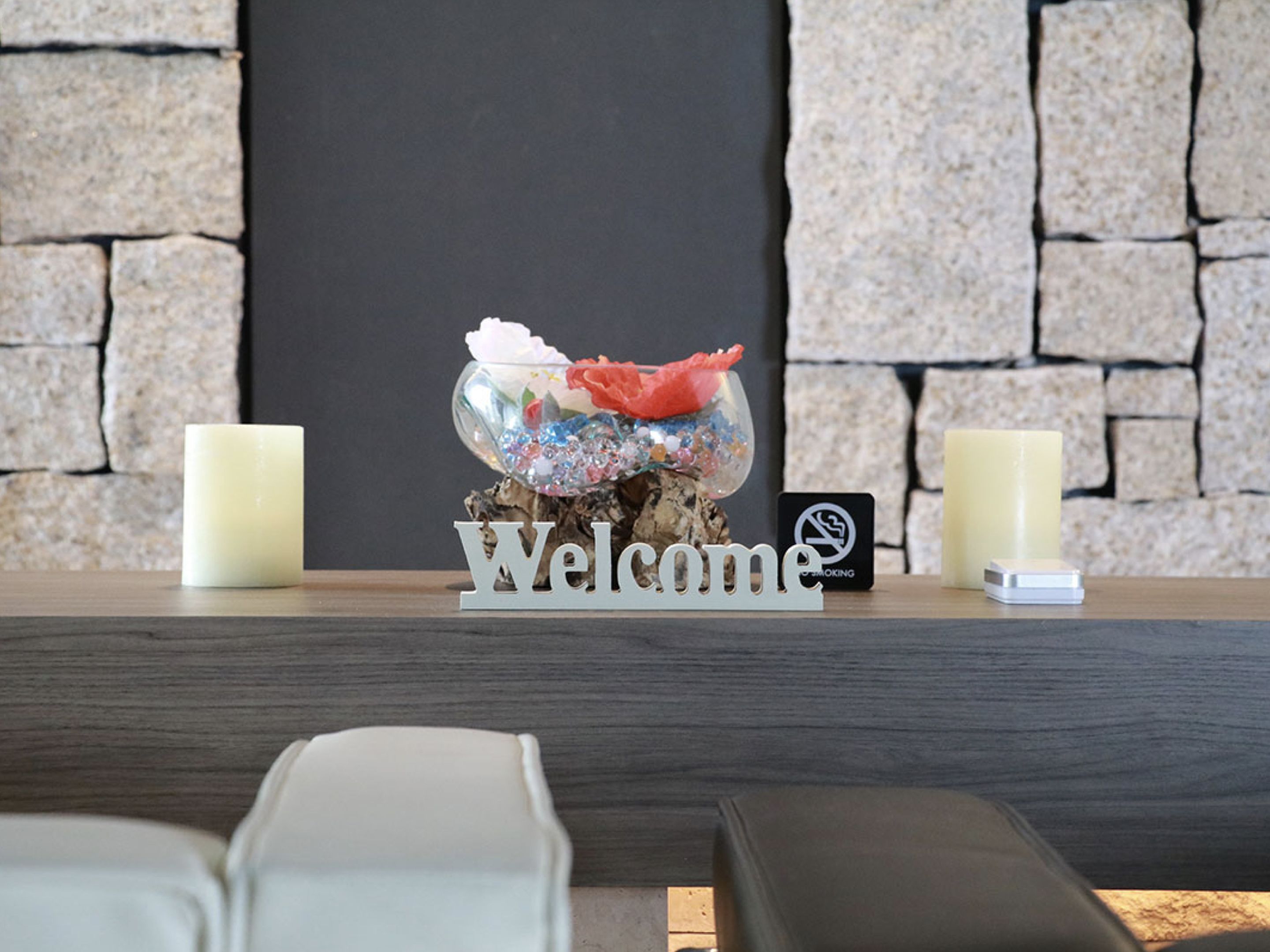 Un área de recepción con un cartel de 'Welcome', dos velas, un cuenco de vidrio decorativo con flores y un letrero de prohibido fumar.