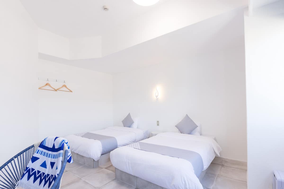Une chambre d’hôtel avec deux lits, une chaise, une lampe fixée au mur et une bouche d’aération au plafond.