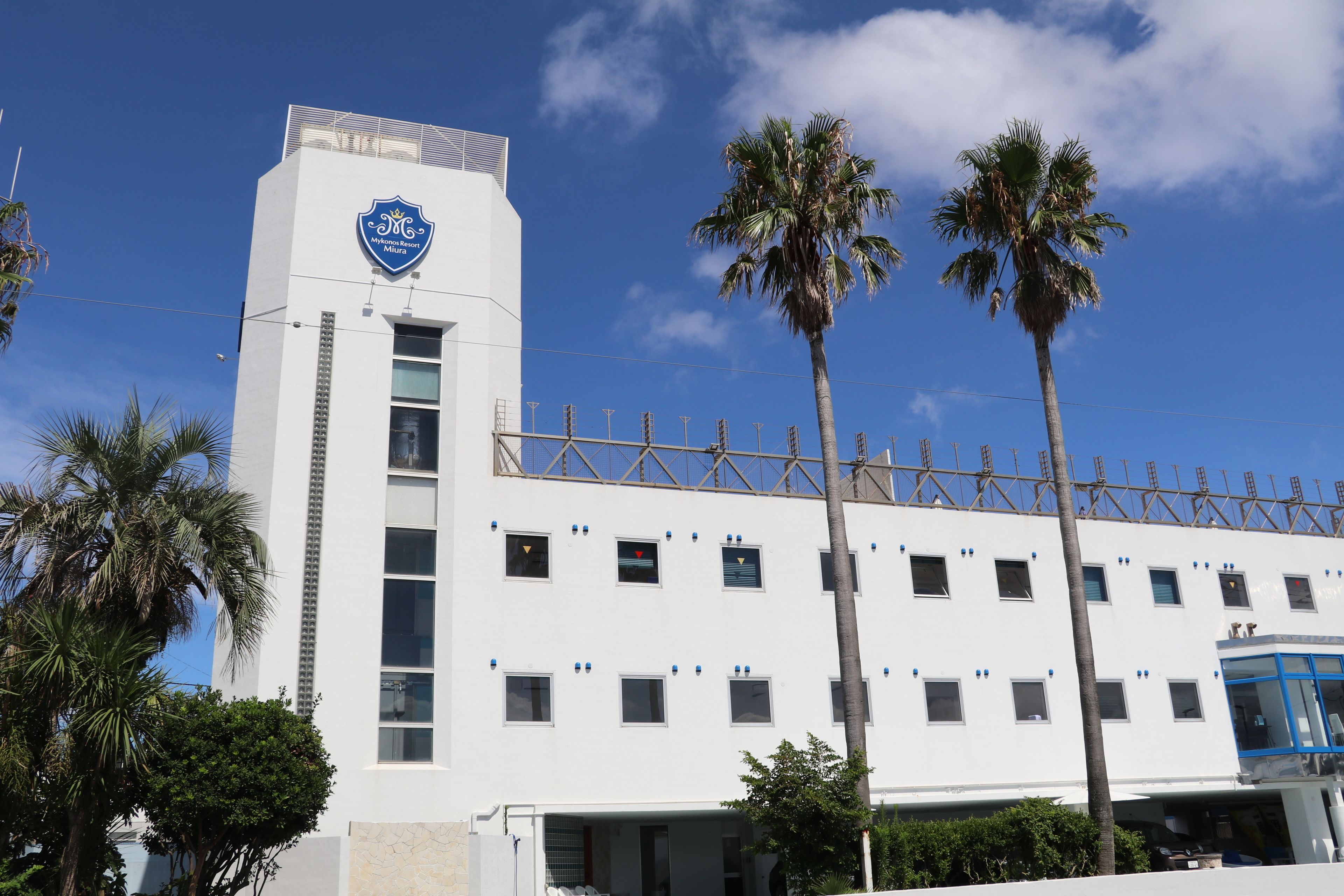 A white building with a blue logo on the top, surrounded by palm trees.