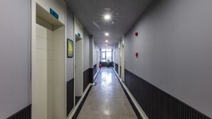 Un long couloir étroit, doté d’un sol carrelé, bordé de murs percés de portes et muni d’un détecteur d’incendie sur le côté droit.