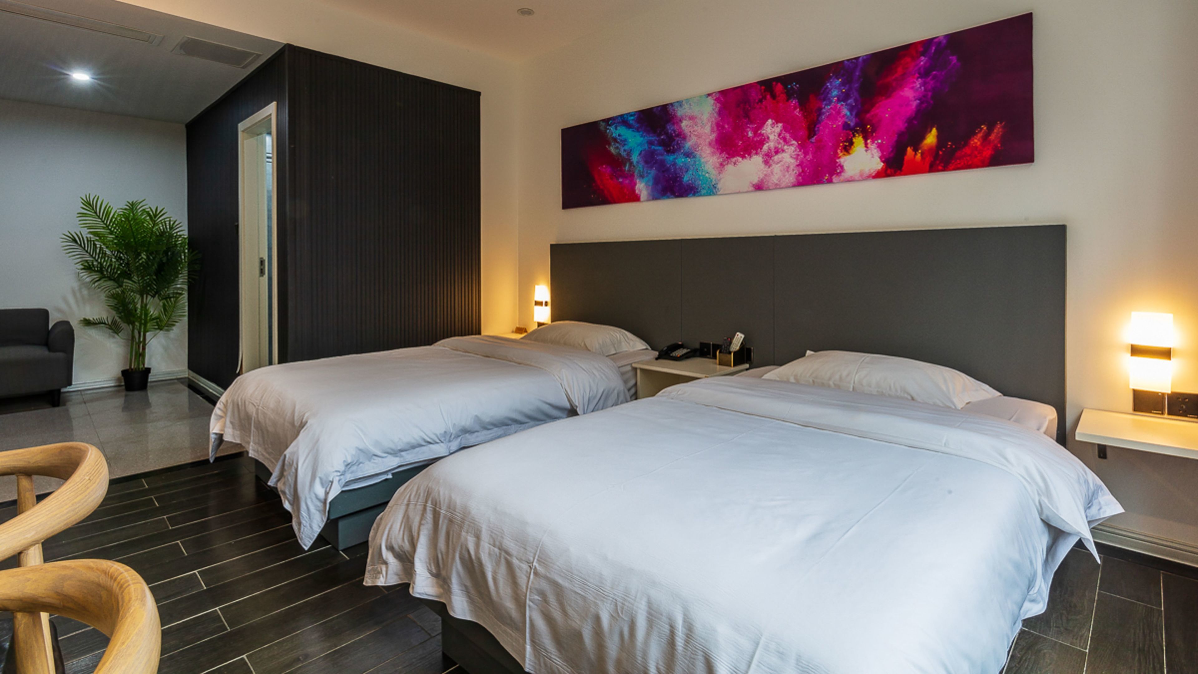 Une chambre d’hôtel avec deux lits, une grande peinture abstraite, une plante en pot et un fauteuil moderne.