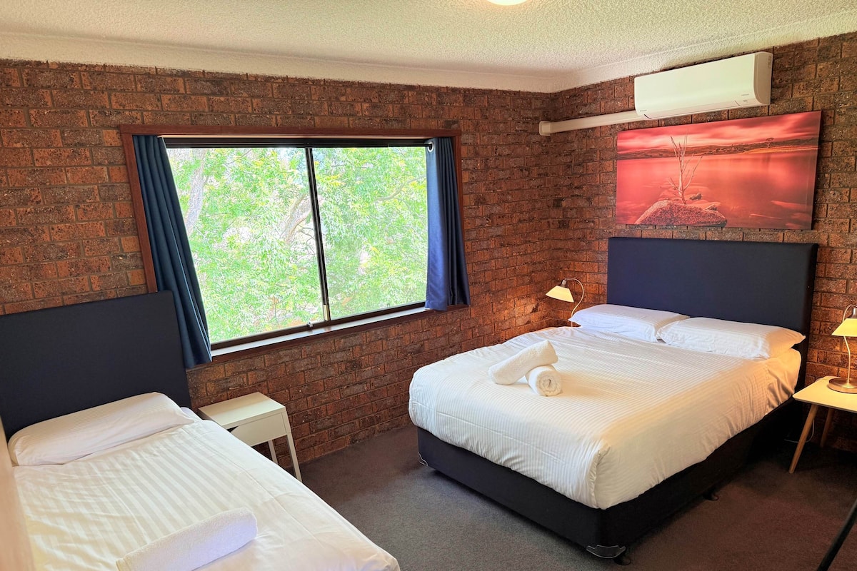 Habitación de hotel con dos camas, una pared de ladrillo, un ventanal con cortinas, un equipo de aire acondicionado y un cuadro en la pared.