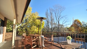 A covered outdoor patio with wooden furniture, a metal heater, and a railing overlooking a body of water.