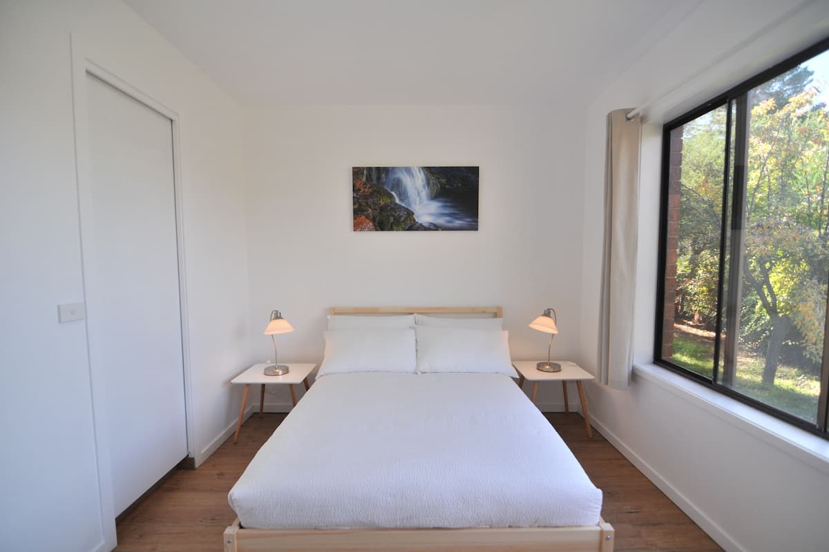 Un dormitorio con una cama grande, dos mesitas de noche con lámparas, un cuadro de una cascada y una ventana con vistas a árboles.